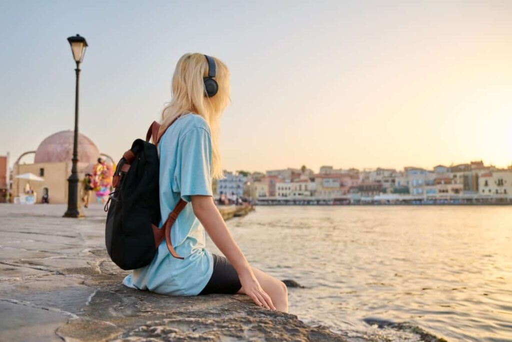 Happy young teenage female in headphones with backpack enjoying old tourist European city, seascape, copy space. Tourism, Greece Crete Chania, summer vacation, youth, rest, nature, sunset